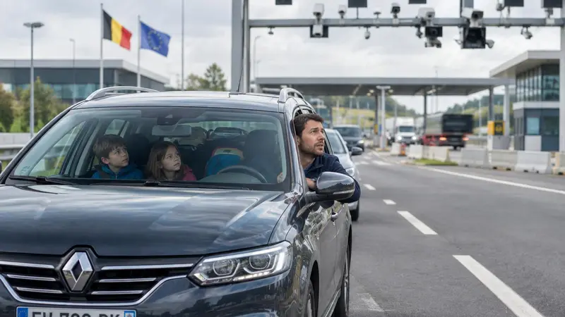 Familie im Auto an einer Grenzstelle mit Kamerabrücke, im Hintergrund wehen die belgische und die europäische Flagge