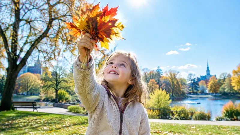 Ein Mädchen hält einen Strauß gelber, oranger und roter Herbstblätter in die Sonne, das Licht scheint durch die Blätter.