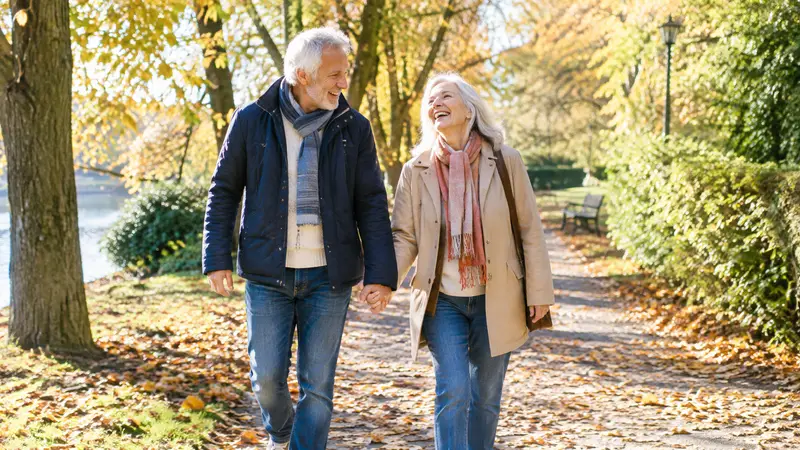 Ein älteres Paar spaziert lachend Hand in Hand durch einen Park mit bunt gefärbten Herbstbäumen und Laub auf dem Weg.