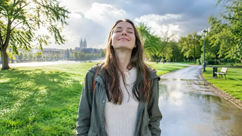 Eine junge Frau steht nach einem Regenschauer auf einem nassen Parkweg, schließt die Augen und atmet lächelnd tief ein.