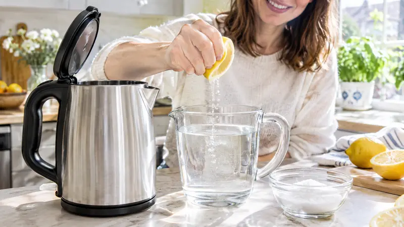 Eine Frau presst eine Zitrone über einem Messbecher mit Wasser aus, daneben ein Wasserkocher und eine Schale Zitronensäure